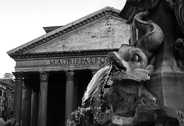 Close view of the fountain in the square: a sculpted fish spouts water, with the Pantheon pediment and part of the Agrippa inscription behind
