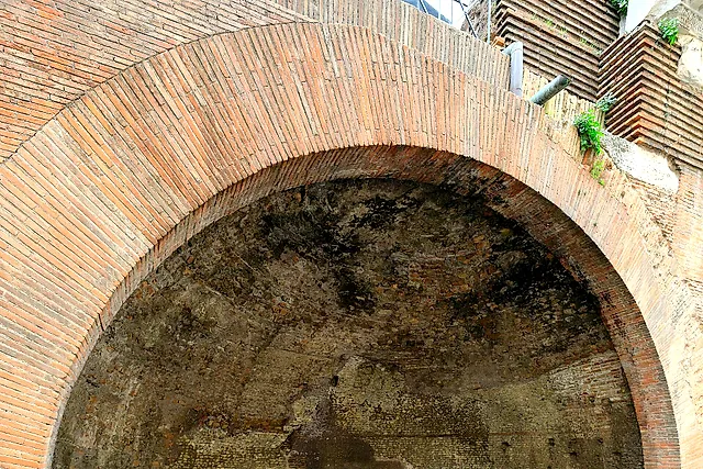 A brick arch framing a niche in the outer wall, with a stone base and loose blocks of marble on the paving